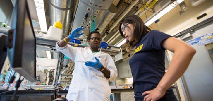 A person in lab research gear stands next to a person with a U-M t-shirt as they look at a screen